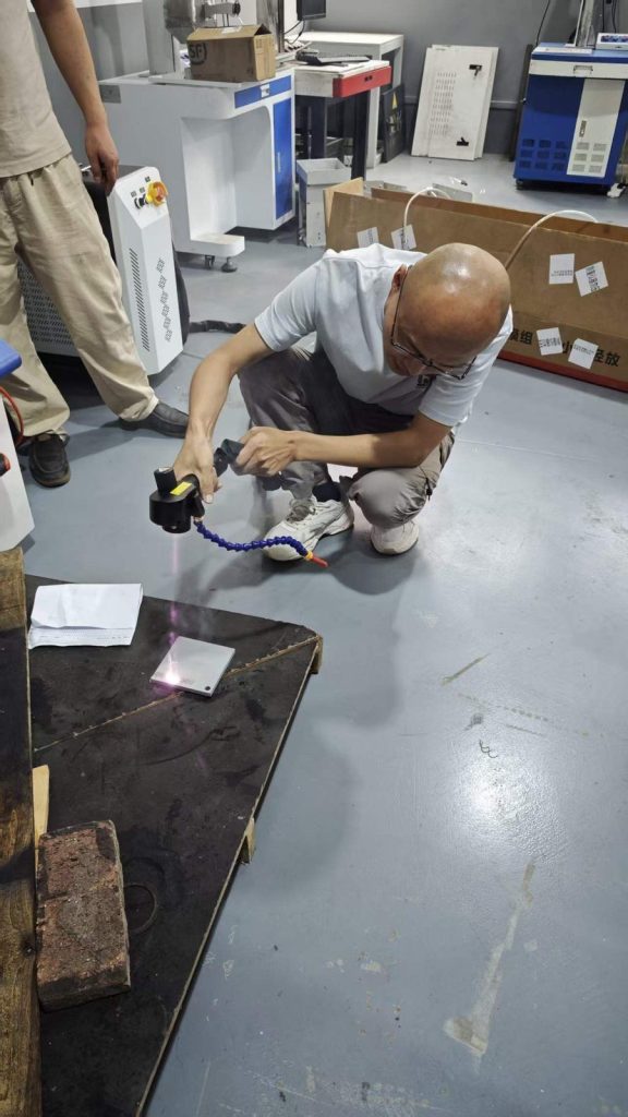 Handheld pulsed laser cleaning machine positioned for routine maintenance checks and sample-test review on a metal panel
